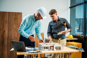 A Caucasian male engineer and an Italian male building contractor are planning and talking at a desk, reviewing house plans and a model of a house with wooden textures for renovation.