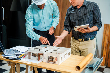 A Caucasian male engineer and an Italian male building contractor are planning and talking at a desk, reviewing house plans and a model of a house with wooden textures for renovation.