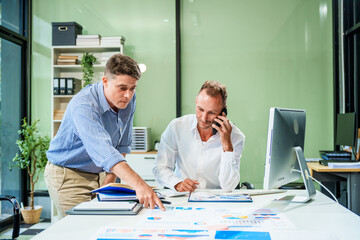 Obraz premium An Italian male and a Caucasian male sit at business desk in office meeting room, discussing financial terms, investment strategies, managing funds with computer monitor displaying relevant data.