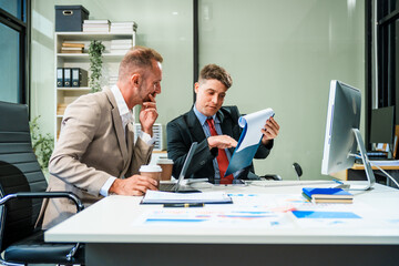 An Italian male and a Caucasian male, both in formal suits, sit at a business desk in an office meeting room with a desktop monitor and laptop, discussing business finance and investment strategies.