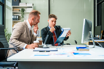 An Italian male and a Caucasian male, both in formal suits, sit at a business desk in an office...