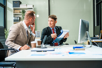 An Italian male and a Caucasian male, both in formal suits, sit at a business desk in an office...