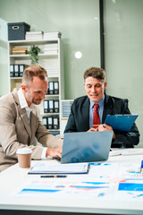 An Italian male and a Caucasian male, both in formal suits, sit at a business desk in an office...