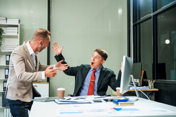An Italian male and a Caucasian male, both in formal suits, sit at a business desk in an office meeting room with a desktop monitor and laptop, discussing business finance and investment strategies.