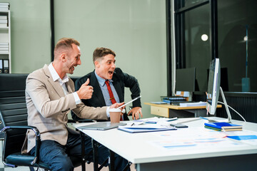 An Italian male and a Caucasian male, both in formal suits, sit at a business desk in an office...