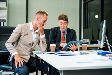 An Italian male and a Caucasian male, both in formal suits, sit at a business desk in an office meeting room with a desktop monitor and laptop, discussing business finance and investment strategies.