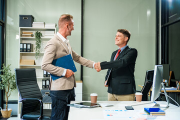 An Italian male and a Caucasian male, both in formal suits, sit at a business desk in an office...