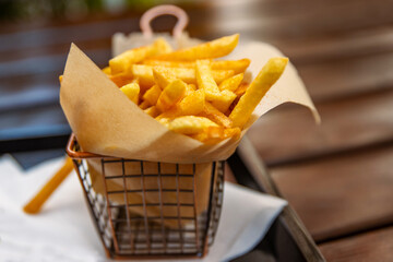 Close up view of golden, crispy French fries served in a stylish wire basket lined with brown parchment paper. Ideal for food photography, restaurant menus, and culinary promotions. Selective focus