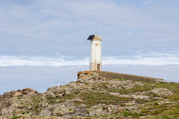 Showcases an old, Roncudo in Corme, Spain weathered lighthouse standing on a rocky coast under a partly cloudy blue sky, highlighting the serene coastal landscape and historic structure.