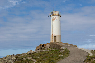 Showcases an old, Roncudo in Corme, Spain weathered lighthouse standing on a rocky coast under a partly cloudy blue sky, highlighting the serene coastal landscape and historic structure.