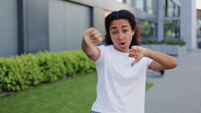Angry woman making thumb down gesture while complaining to camera and waving no with index finger. Irritated female giving negative answer to proposition of coworking or partnership at empty street.