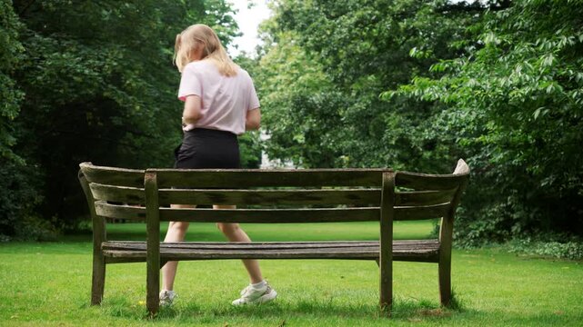 young woman with coffee in hand sits on bench in park and enjoys drink. Back view of girl who approaches wooden bench in sunbeams and sits on it.