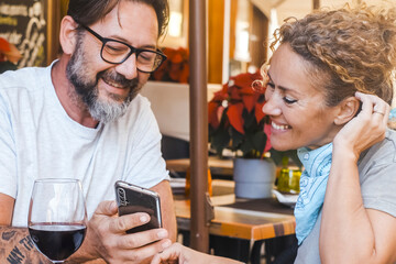 Modern adult couple sitting at table restaurant watching smartphone to leave a review feedback after lunch. Choosing the best pizzeria before eat reading comments on line on website on the phone