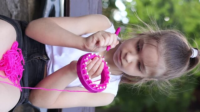 A girl knits on a loom with pink threads while sitting on a bench in a summer park