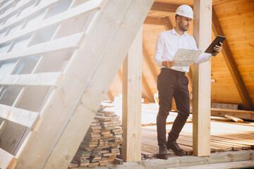 Man builder. Foreman studies blueprints with tablet. Construction company. Architect in unfinished building. Builder in wooden house under construction. Man engineer from construction team