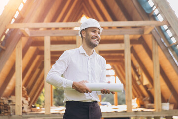 Construction engineer in white helmet. Developer with construction documentation on the background of wooden building under construction. Portrait of smiling architect near eco friendly house.