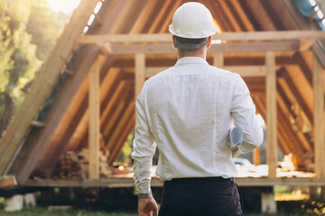 Rear view of construction engineer in white helmet. Developer with documentation on the background of wooden modular building under construction. Portrait of smiling architect near eco friendly house