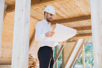 Fototapeta premium Construction engineer in white helmet. Developer with construction documentation on the background of wooden modular building under construction. Portrait of smiling architect near eco friendly house.