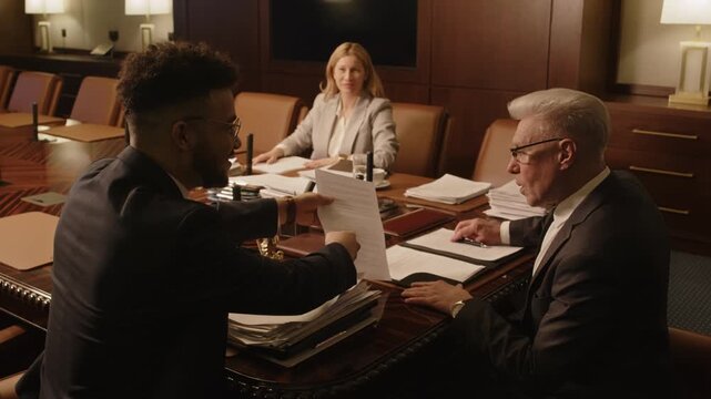 Zoom out shot of three lawyers sitting at desk in meeting room and celebrating successfully closed case senior man giving young male colleague high five, woman clapping hands happily