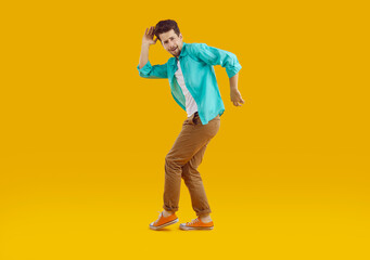 Full length portrait of brunet young bearded crazy cheerful man in turquoise shirt and brown pants funny dancing on yellow background. Disco, dance, banner for advertisement and marketing concept.
