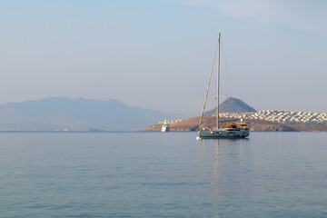 White yacht with lowered sails at sea on a sunny day against the background of mountains, a peninsula with houses early in the morning