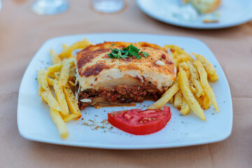 A slice of Greek moussaka with French fries and tomato on a white plate