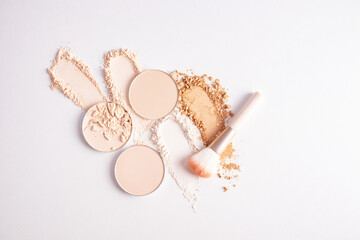 several round blocks of compact powder in light tones and swatches of loose powder in different tones on a white background with a makeup brush
