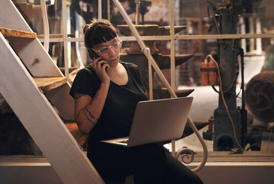 Woman, laptop and phone call or talk in factory, foundry and inventory for raw materials. Professional, quality control and browsing for service worker for machines, email and booking conversation