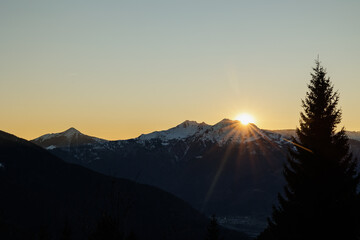 sole che tramonta dietro le montagne scure e innevate, in un cielo sereno e di colore misto giallo azzurro, in inverno