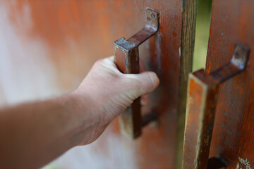 A hand is opening a door with a rusty handle. The door is wooden and has a metal hinge