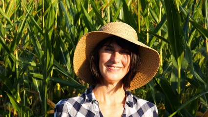 Happy smiling female farmer looks into camera standing near corn field. Portrait of adult beautiful agronomist in straw hat with maize meadow at background. Agricultural business concept