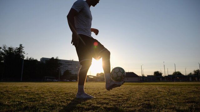 Professional footballer juggling soccer ball on stadium at sunset. Young man kicking ball at green field. Sportsman practicing tricks at meadow with sunlight at background. Freestyle football concept