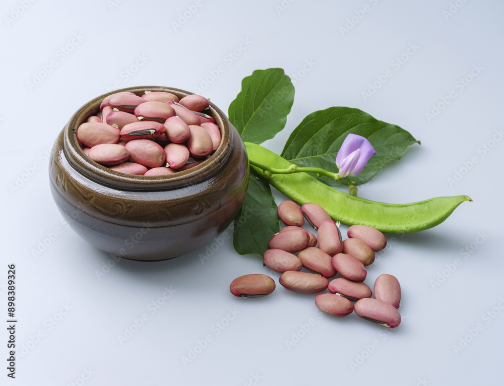 Sticker Close-up of raw red horse bean seeds on small jar with green pod, leaves and pink flower on white floor, South Korea
