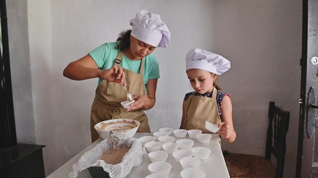 Family baking together indoors. Mother and daughter making cupcakes in the kitchen, wearing aprons and chef hats.