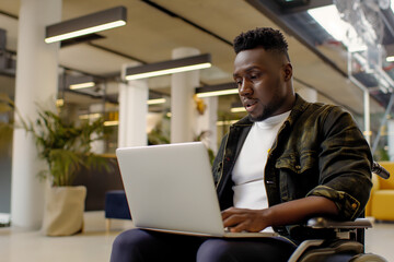 Young black man in a wheelchair, working on a laptop, in a modern office, surrounded by plants and furniture,  Focused man with a disability, using a laptop, in a bright and modern office environment
