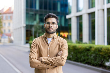 Confident young man with glasses standing outside modern office building on city street. Wearing casual brown shirt, arms crossed, looking at camera.