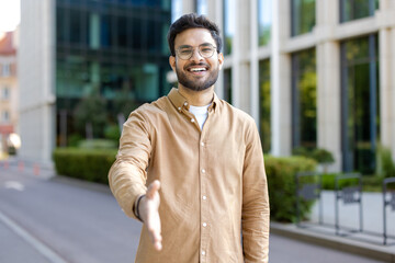 Confident smiling man extending hand for handshake outside modern building. Person wearing glasses and a casual shirt, standing on urban street. Welcoming gesture in professional or casual encounter