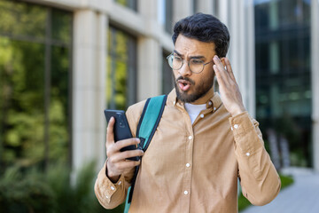 Young man in glasses standing outside looking shocked while checking his smartphone, expressing surprise and disbelief. Professional setting with modern building and greenery in background.