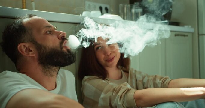 A guy with a beard in a white T-shirt smokes an electronic cigarette with his brunette girlfriend while sitting on the floor in the kitchen in the morning