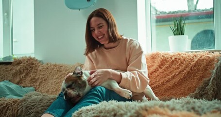 Happy brunette girl holds her pet dog in her arms and strokes it while sitting on a soft sofa in a modern apartment
