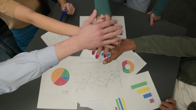 Top view paperwork on table hands stacking in pile diverse business team men women coworkers girls guys put arms together unity gesture union support motivation teamwork partnership win celebration