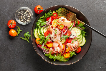 Salad with fresh vegetables and jamon in a black bowl on the table. Top view