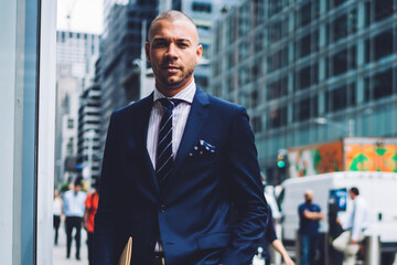 Portrait of confident young entrepreneur dressed in elegant suit holding hand in pocket while looking at camera.Serious businessman with folder for documents standing in urban setting of downtown