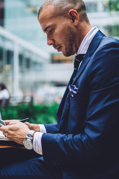 Pensive businessman in formal wear sitting in urban setting and chatting online on smartphone device using public 4G internet connection.Young entrepreneur in suit reading notification on cellula