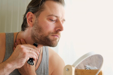 Reflection of a young bearded man shaving his beard holding electric shaver in bathroom. Handsome...