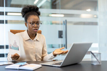 Frustrated businesswoman analyzing financial report while sitting at office desk with laptop. Professional woman experiencing work stress or confusion in modern corporate environment