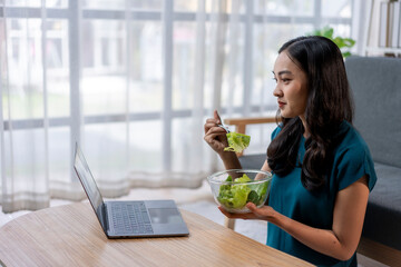 A woman is eating a salad while looking at a laptop