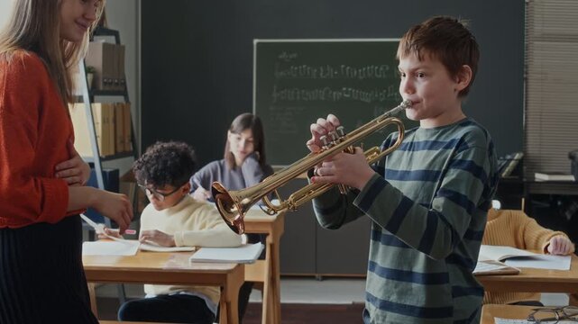 School boy learning to play trumpet while smiling female teacher watching him, children completing task in background sitting at desks in music class
