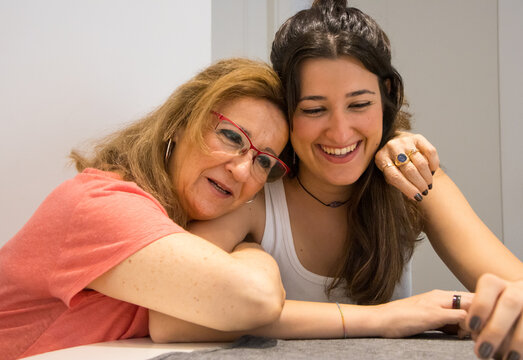 Attractive mother and her young daughter chatting quietly sitting at the table at home hugging and sharing a family moment on the weekend wearing different jewelry.
