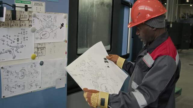 Side medium shot of Black male engineer in protective overalls and hardhat looking at pictorial blueprint of machine detail in heavy machinery plant